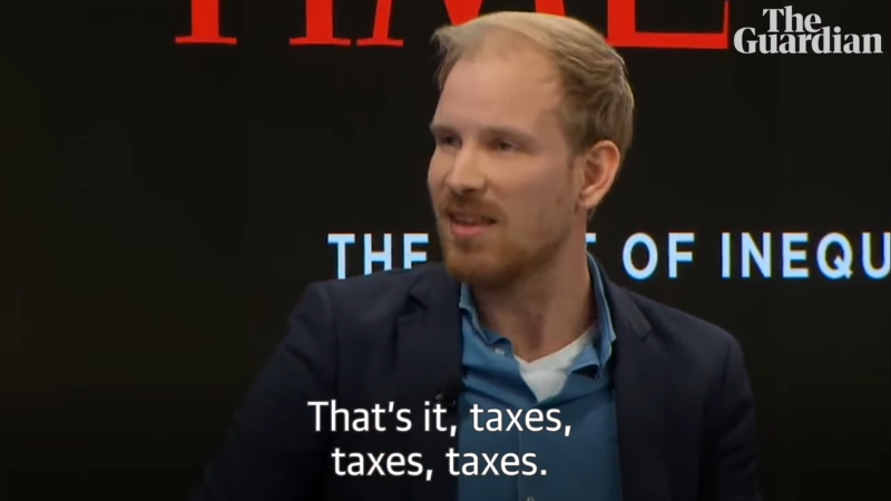 A man, seated and facing slightly to the left of the camera. He has light-colored hair and a beard, and he is wearing a dark blazer over a light blue shirt and a white undershirt. His expression is engaged and he seems to be in the middle of making a point.