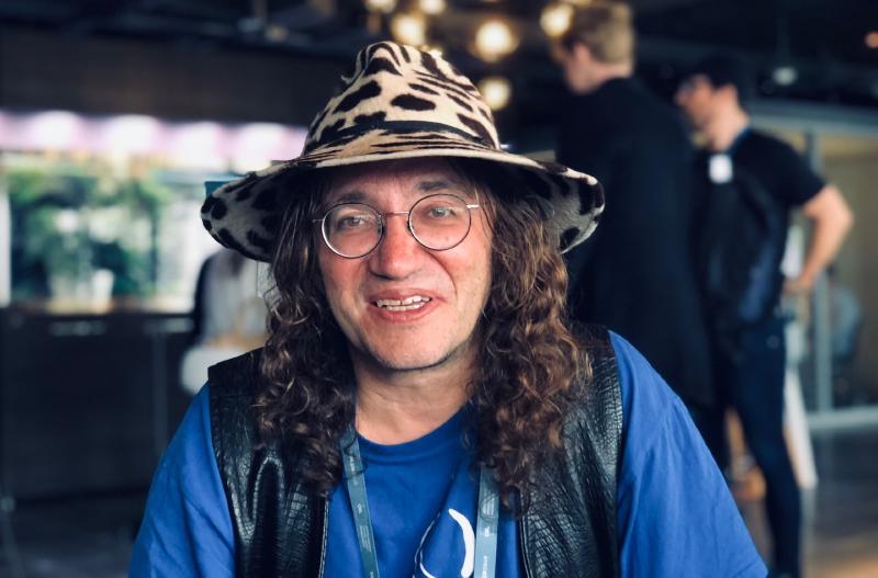 A man in his fifthies, with long curly hairs, round glasses and a bizarre wide-brimmed hat, sporting a cow-print design.
