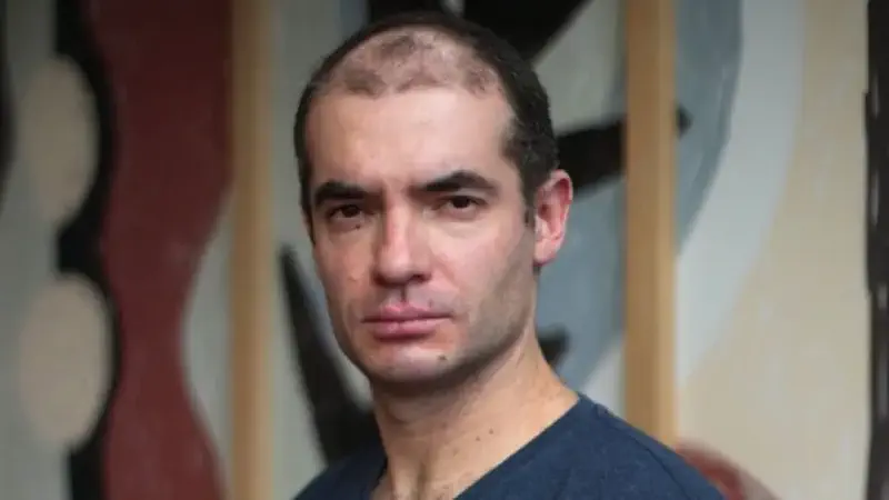 A man in his 30s or 40s, with a receding hairline and closely cropped hair. He has a neutral expression, prominent eyebrows, and is wearing a dark blue V-neck shirt. 